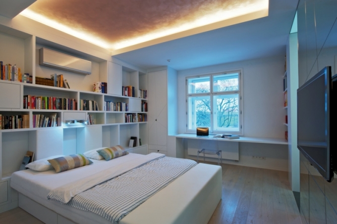 Ložnice má spoustu úložných a odkládacích prostor - skříně, zásuvky pod postelí, policový systém integrovaný do hlavní stěny i jednoduchý stůl pod oknem. K útulné atmosféře přispívá po obvodu místnosti snížený strop se zabudovaným nepřímým osvětlením i masivní podlaha z běleného dubu
