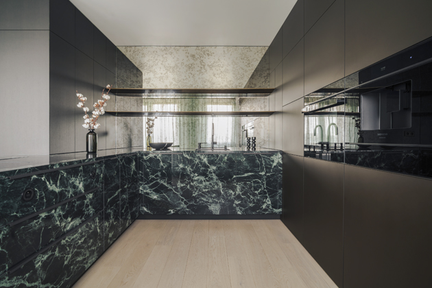 Zelený mramor se autoři rozhodli využít také pro část spodních kuchyňských dvířek. Jedná se o velkoformátový obklad, který byl nainstalován na truhlářské prvky. To pochopitelně vyžadovalo precizní technickou přípravu a důkladné promyšlení jednotlivých detailů. Jen díky tomu výsledek působí vizuálně čistě a je také plně funkční. V prostřední části kuchyně pak pozornost poutá stěna obložená „stařeným“ zrcadlem.